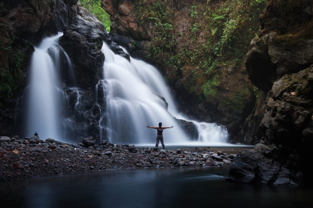 Person standing with open arms in front of a peaceful waterfall, symbolizing healing, self-discovery, and reconnecting with yourself.